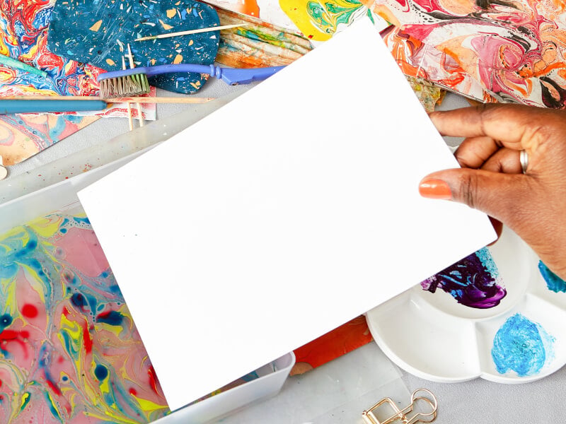 A person is dipping a piece of paper into water marbled with inks